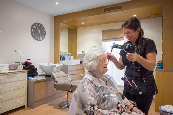 A resident sitting in a salon chair while a hairdresser styles her hair with professional tools and products. Available for all our Anchor residents.
